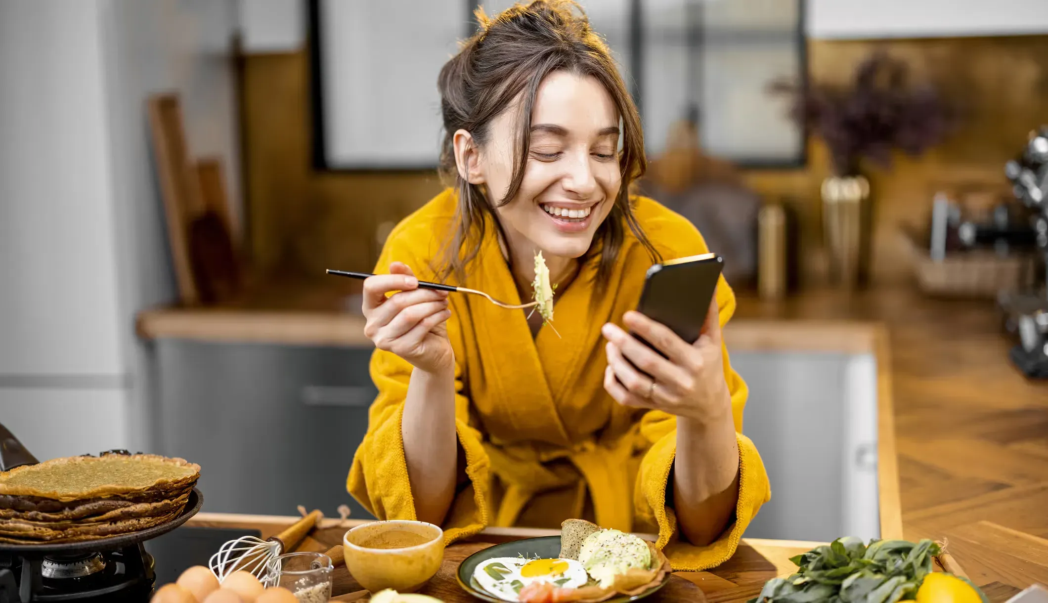 A woman in a yellow robe eating breakfast in her kitchen while she looks at her phone